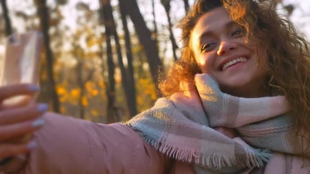Portrait of caucasian curly-haired woman sitting on bench and making selfie-photos using smartphone in autumnal park. - Video