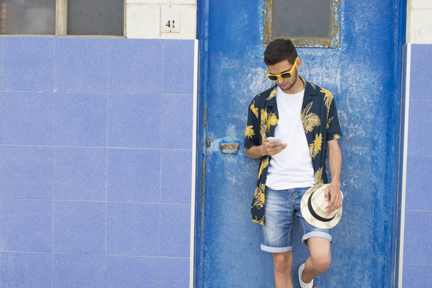 güneş gözlüğü ve mavi duvar üstünde hareket eden telefon ile rahat stil ile genç adam - Fotoğraf, Görsel