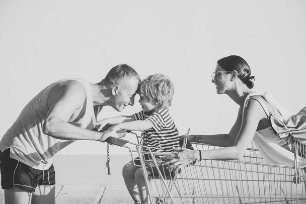 Family shopping concept. Happy parents go shopping with cute son. Couple pushing shopping cart with children in. Mother and father on smiling faces walks with cart on sunny day, white background. - Φωτογραφία, εικόνα
