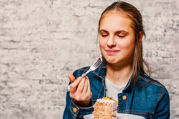 portrait of young teenager brunette girl with long hair eating slice cake dessert on gray wall background - Photo, Image