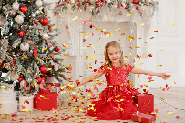 A little girl in a red dress catches confetti on the background of the New Year tree - Fotó, kép