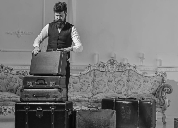 Man, traveller with beard and mustache packing luggage before trip, luxury interior background. Luggage and travelling concept. Macho elegant on thoughtful face standing near pile of vintage suitcase - Zdjęcie, obraz