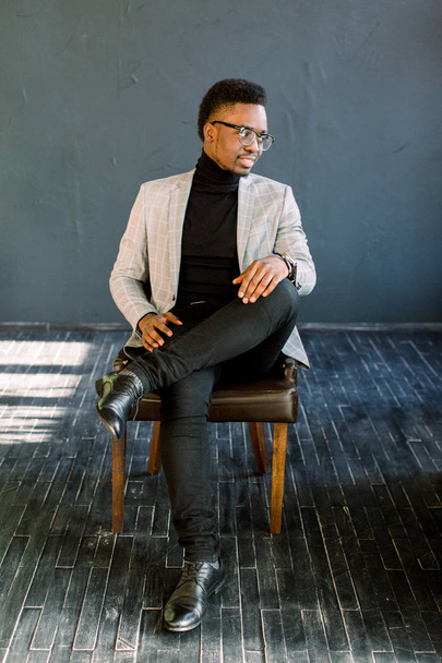 Attractive handsome happy smiling African American businessman wearing a light jacket and glasses sitting on a leather chair on a dark background - Foto, imagen