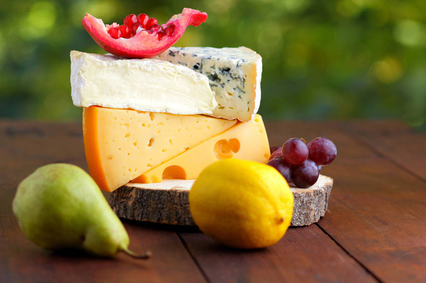 Camembert, brie, hard cheese and grapes on a wooden stand. Pieces of different cheeses, pear and lemon on blurred background. Dorblu, soft cheese and pomegranate on boards. Cheese and fruit - Photo, Image