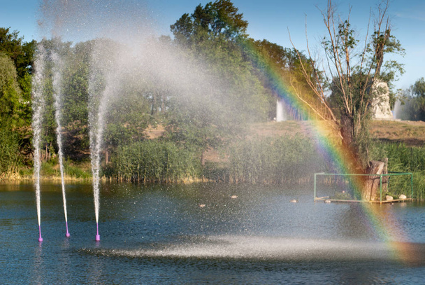Little rainbow from the water fountain lake, city park pond, water jet beats up against the background of green trees, summer weekend rest - Photo, Image