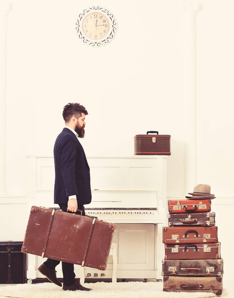 Butler and service concept. Macho attractive, elegant carries vintage suitcases, side view. Man with beard and mustache wearing classic suit delivers luggage, luxury white interior background - Fotografie, Obrázek