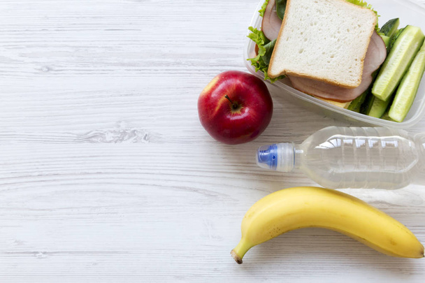 Caja de almuerzo saludable con sándwich, frutas y botella de agua en la mesa de madera blanca, vista superior. Desde arriba, plano, sobre la cabeza. Copiar espacio
. - Foto, Imagen