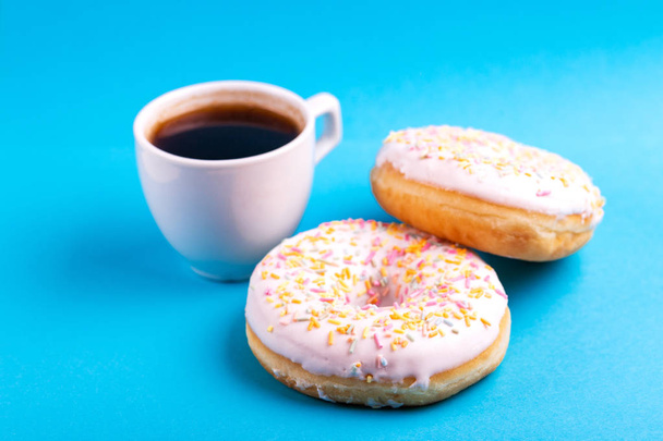 Two delicious vanilla donuts with sprinkle and cup of coffee on bright blue background. Unhealthy, but tasty sweets. Copy space. Horizontal view - Foto, immagini