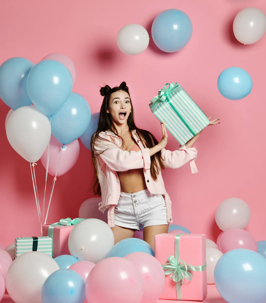 Portrait of young woman in birthday party holding gift box with gift present on pastel pink background with colorful air baloons. - Foto, afbeelding