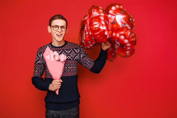 A man wearing glasses with a bunch of flowers and heart-shaped balloons on a red background - Photo, Image