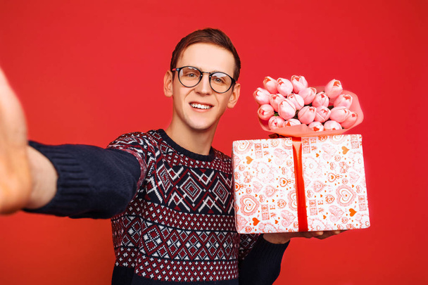 A man in glasses, with a gift in his hands and a bouquet of flowers, photographs himself on a smartphone against a red background. Valentine's Day - Foto, Imagen