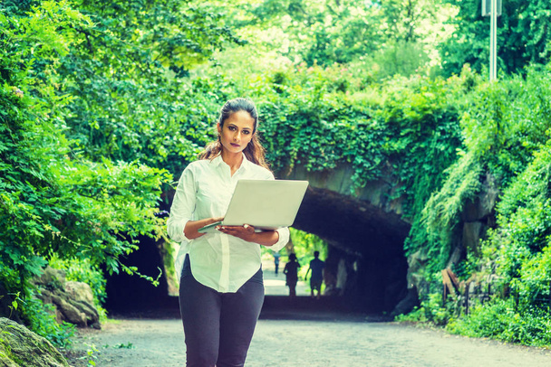 junge ostindische Amerikanerin auf Reisen, arbeitet in New York, trägt weißes Hemd, schwarze Hose, hält Laptop in der Hand, geht im Central Park spazieren. Straßenbrücke mit grünen Blättern im Hintergrund - Foto, Bild