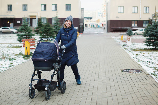 Pretty woman in blue jacket with a pram on the street in the yard in the fall. Baby carriage outdoor/ - Fotó, kép