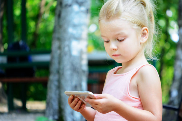 Little girl with blond hair sitting on bench and using smartphone - Fotografie, Obrázek