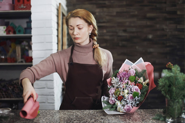 Pretty middle-aged florist woman in apron makes a bouquet in the flower salon for a festive gift for a wedding or anniversary. - Фото, изображение
