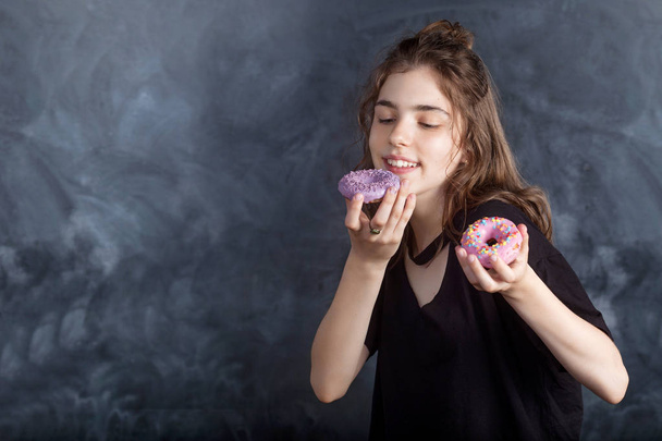 Portrait of joyful girl with donuts on black background. Happy girl holds fresh donuts and looks at them. Good mood, diet concept. - Foto, afbeelding