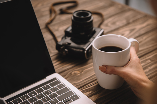 cropped view of freelancer holding cup of coffee at workplace with vintage photo camera and laptop - Photo, Image