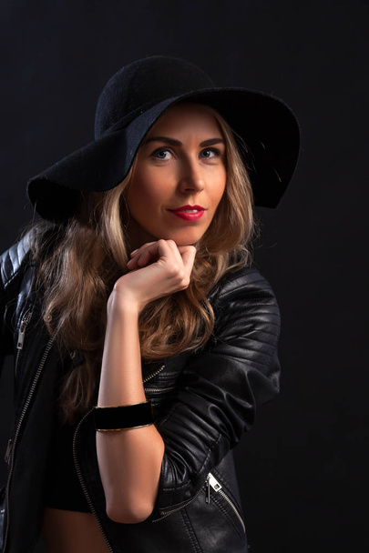 Smiling beautiful young woman in in trendy black T-shirt, orange skirt, leather jacket and black hat  posing with hands on chin. Three quarter length studio shot on  black background - Foto, afbeelding