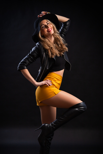 Nahaufnahme Studio Porträt der schönen Glamour sinnliche Frau in trendigen schwarzen T-Shirt, orangefarbenem Rock, Lederjacke und Hut auf schwarzem Hintergrund. gesunde wellige lange Haare.  - Foto, Bild
