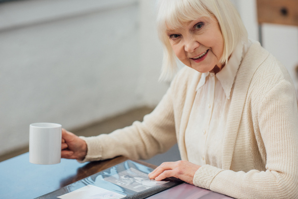 Seniorin sitzt mit Fotoalbum am Tisch und schaut in Kamera, während sie zu Hause Kaffee trinkt - Foto, Bild