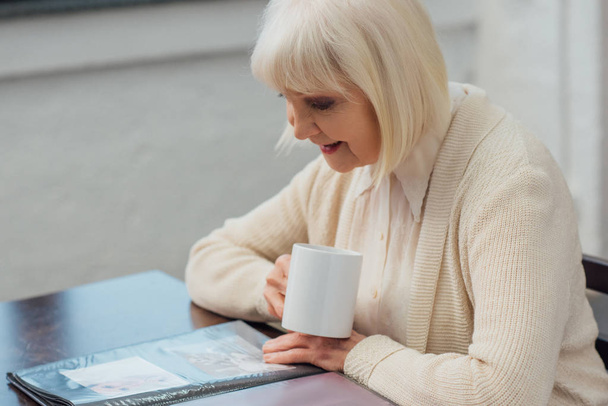 Senior kobieta siedzi przy stole i patrząc na album fotograficzny podczas picia kawy w domu  - Zdjęcie, obraz