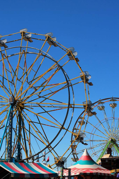 Два колеса обозрения в силуэте против ярко-голубого неба с красочными тентами на полпути в нижней части вертикального вида
 - Фото, изображение