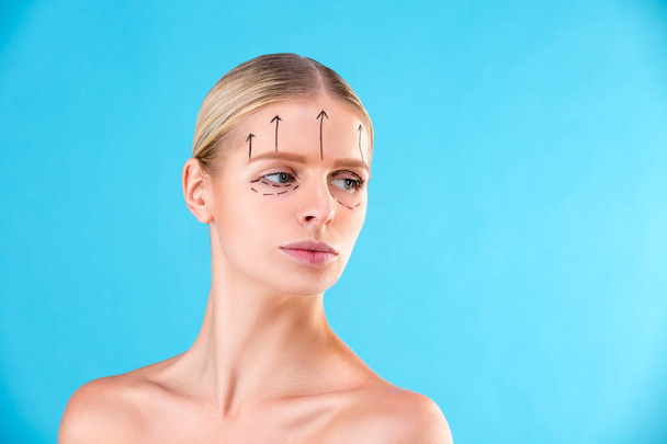 Beautiful Woman Face. Beauty Portrait of young woman blond, painted lines on the face for cosmetic surgery on blue background. Cosmetic surgeon blepharoplasty. Lifting - Photo, Image