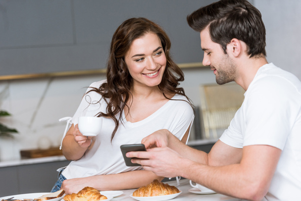 alegre novio sosteniendo teléfono inteligente cerca de novia con taza de café
 - Foto, imagen