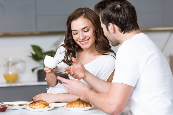 smiling woman looking at smartphone in hand of boyfriend in kitchen  - Фото, зображення