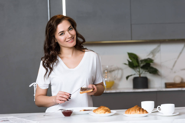 attractive woman smiling while holding knife and toast bread in kitchen  - Photo, Image