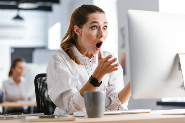 Shocked young woman dressed in shirt sitting at her workplace at the office, looking at computer - Foto, Bild