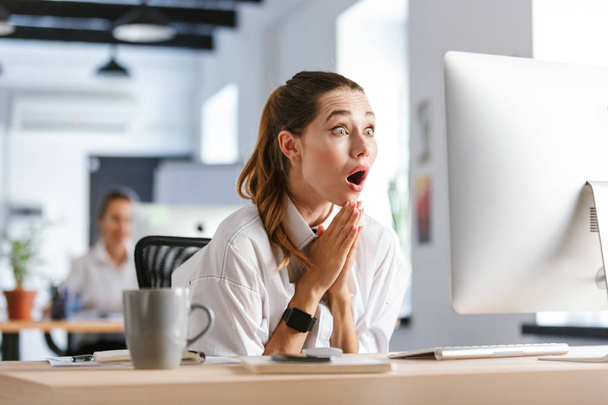 Shocked young businesswoman sitting at her workplace at the office, looking at computer - Foto, Bild