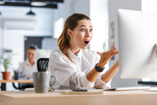 Shocked young businesswoman sitting at her workplace at the office, looking at computer - Foto, Imagem