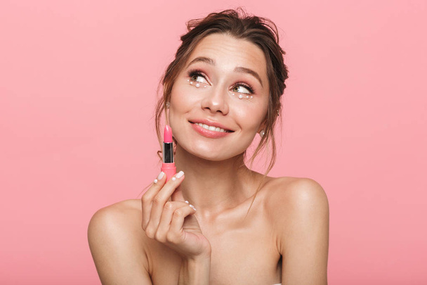 Image of a pretty thinking happy young woman posing isolated over pink wall background holding lipstick. - Foto, Imagem
