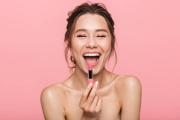 Image of a pretty happy young woman posing isolated over pink wall background holding lipstick. - Photo, image