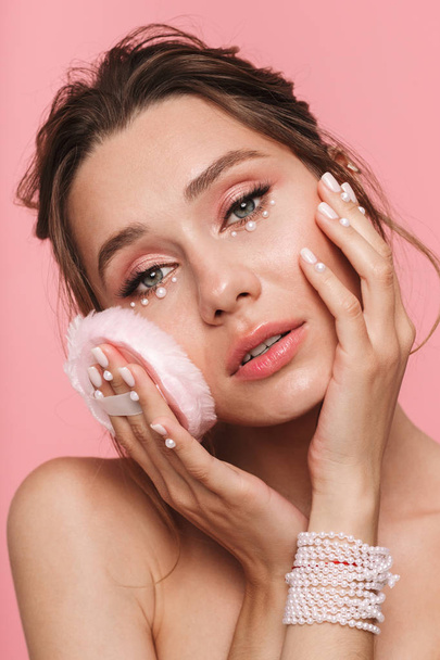 Beauty portrait of an attractive young topless woman using powder puff isolated over pink background, wearing white pearls - 写真・画像