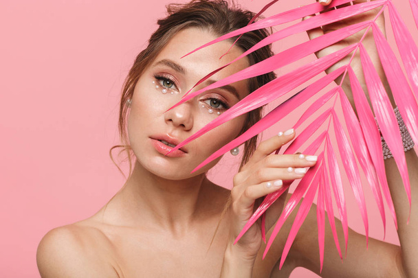 Beauty portrait of an attractive young topless woman posing with pink palm leaves isolated over pink background, wearing white pearls - Foto, imagen