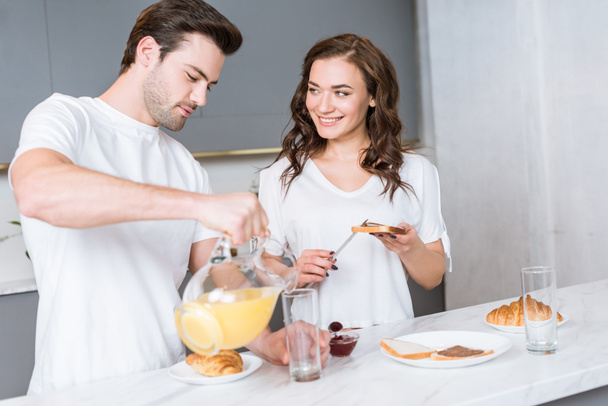 namorada olhando para homem bonito segurando jarro com suco de laranja na cozinha
  - Foto, Imagem