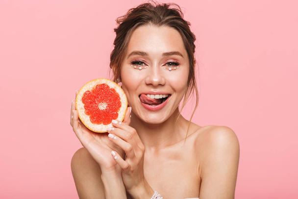 Beauty portrait of an attractive healthy young topless woman posing with grapefruit isolated over pink background, wearing white pearls - Foto, afbeelding