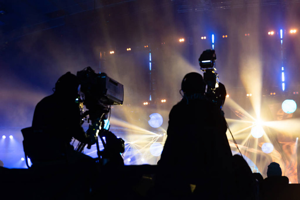 Television broadcast by a cameraman during a concert. Camera with the operator is on the high platform. Silhouettes on the background of colorful beams. - Photo, Image