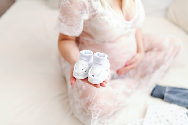 Close up van zwangere vrouw houden baby's schoeisel in de ene hand terwijl zittend op het bed met de benen gekruist. Andere hand op buik. - Foto, afbeelding