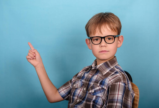a cute European boy with glasses points his finger at an empty background . blank for design and advertising on blue background - Φωτογραφία, εικόνα