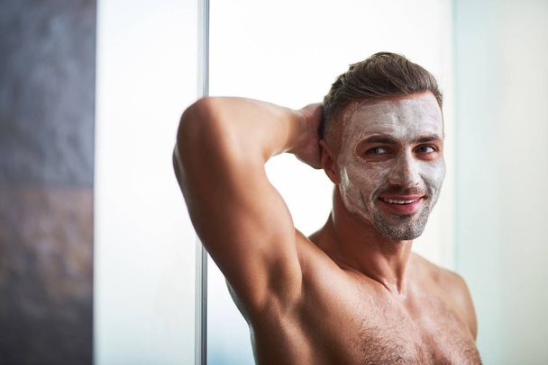 Attractive young man with hand behind his head using facial mask - Fotografie, Obrázek