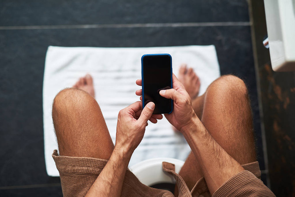 Young gentleman using mobile phone while sitting on toilet bowl - Foto, afbeelding
