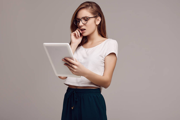 Portrait of cute teenage girl in white shirt and glasses holding digital tablet on gray background. Studio shoot. - Фото, изображение