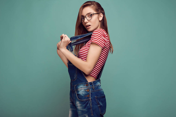 Портрет милой девочки-подростка в джинсовом комбинезоне и очках, позирующих на цветном фоне. Студийные съемки
. - Фото, изображение