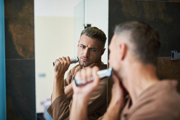 Attractive serious man looking in the mirror and shaving - Foto, Bild