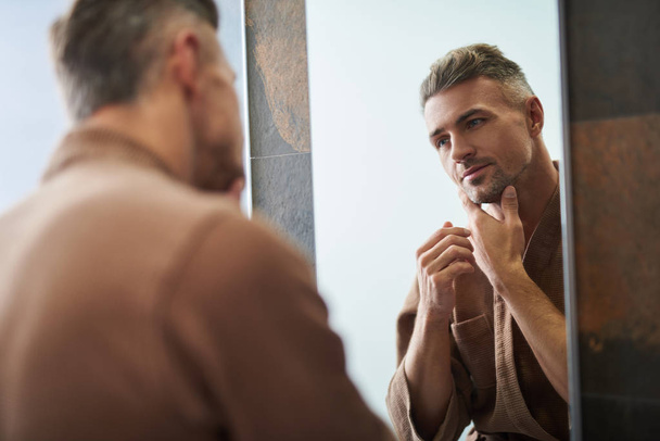Handsome unshaven man looking in the mirror at home bathroom - Фото, изображение
