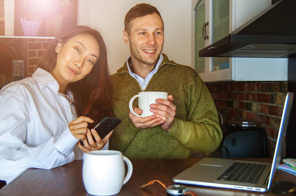 Les jeunes couples à la maison près de l'ordinateur portable boivent du café et rient. Heureux couple international faire un achat en ligne
. - Photo, image