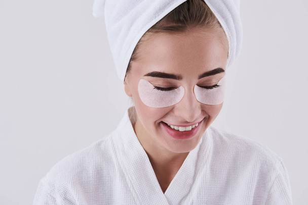 Joyful young lady with towel on head using under-eye patches - Fotografie, Obrázek